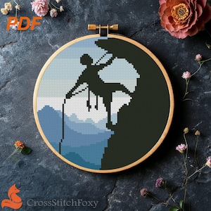 Op de afbeelding: Een kruissteekpatroon in een houten ring toont een bergbeklimmer in silhouet tegen een blauw-witte berglandschap. Het ontwerp is omlijst door een houten ring en omgeven door bloemen. De tekst "CrossStitchFoxy" staat onderaan.