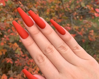 May include: Close-up of a hand with long, orange-red nails. The nails are a matte finish and are all the same length. The hand is against a background of fall foliage.