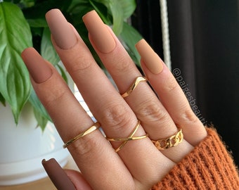May include: Close-up of a hand with long, nude-colored, matte nails. The hand is wearing several gold rings, including a chain link ring. The hand is resting on a brown sweater. The text "@paidesbeauty" is visible in the background.