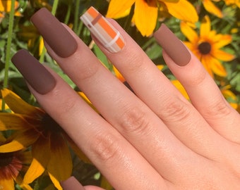 May include: Close-up of a hand with long, square-shaped nails painted in a matte brown color. The ring finger nail features a plaid design with orange, brown, and white stripes.