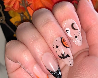 May include: Close-up of a hand with five nails painted with Halloween-themed designs. The nails are painted with a clear base and feature black and orange accents. The designs include a ghost, a crescent moon, a bat, a pumpkin, and stars. The text "@paigesbeauti" is visible on the hand.