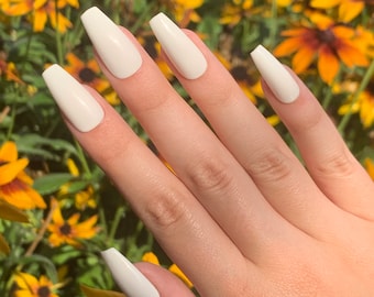May include: A hand model wearing a set of long, white, coffin-shaped press-on nails. The nails are a creamy white color and have a glossy finish.