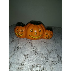 May include: Three ceramic jack-o'-lantern candle holders. The center jack-o'-lantern is the largest and has a wide, toothy grin. The two smaller jack-o'-lanterns are on either side of the center one and have smaller grins.