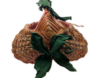 May include: A woven basket with a diamond-shaped top and green paper leaves. The basket is made of light brown and peach-colored materials. The basket has a handle and is decorated with green paper leaves.