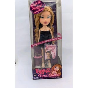 May include: A Bratz Meygan fashion doll in a clear plastic box. The doll has long, light brown hair and is wearing a black dress. The box has text that reads "Collect All Meygan Fashion Heads!" and "Bratz Head Gamez!"