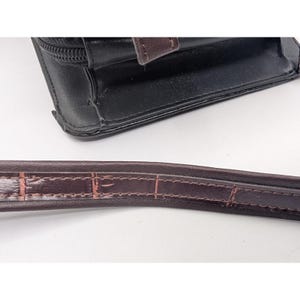 K&ouml;nnte beinhalten: Schwarze Ledergeldb&ouml;rse mit braunem Riemen. Die Geldb&ouml;rse hat einen Rei&szlig;verschluss und einen braunen Lederakzent. Der Riemen ist dunkelbraun mit einem strukturierten Muster.