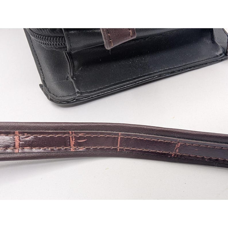 K&ouml;nnte beinhalten: Schwarze Ledergeldb&ouml;rse mit braunem Riemen. Die Geldb&ouml;rse hat einen Rei&szlig;verschluss und einen braunen Lederakzent. Der Riemen ist dunkelbraun mit einem strukturierten Muster.