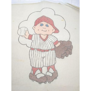 Può includere: Un disegno a punto croce a tema baseball con un giocatore di baseball dei cartoni animati con un berretto rosso e un'uniforme a righe. Il giocatore tiene in mano una palla da baseball e un guanto, su uno sfondo a forma di nuvola. Il disegno è su un tessuto a rete bianco.