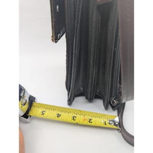 K&ouml;nnte beinhalten: Eine braune Ledergeldb&ouml;rse mit mehreren Rei&szlig;verschlussf&auml;chern. Die Geldb&ouml;rse wird mit einem Ma&szlig;band gemessen, das eine Breite von ca. 13 cm anzeigt. Die Geldb&ouml;rse hat einen Schulterriemen und eine strukturierte Au&szlig;enseite.