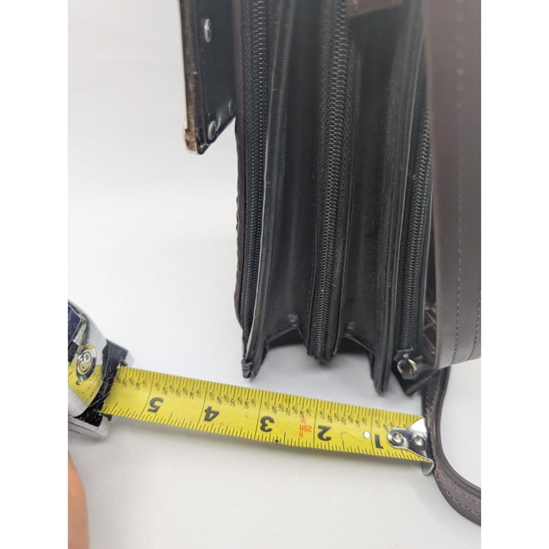 K&ouml;nnte beinhalten: Eine braune Ledergeldb&ouml;rse mit mehreren Rei&szlig;verschlussf&auml;chern. Die Geldb&ouml;rse wird mit einem Ma&szlig;band gemessen, das eine Breite von ca. 13 cm anzeigt. Die Geldb&ouml;rse hat einen Schulterriemen und eine strukturierte Au&szlig;enseite.