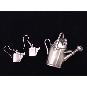 Può includere: Un set di orecchini a forma di annaffiatoio color argento e un ciondolo a forma di annaffiatoio color argento più grande. Gli orecchini sono di tipo pendente con chiusura a gancio. Il ciondolo ha un manico e un beccuccio.