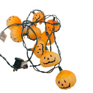 Op de afbeelding: Een slinger van oranje pompoenvormige lampjes met zwarte uitgesneden gezichten. De lampjes zijn geregen aan een groene draad en hebben een standaard elektrische stekker. Perfect voor Halloween-decoraties.