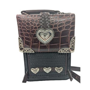 K&ouml;nnte beinhalten: Braune und schwarze Umh&auml;ngetasche mit strukturierter Kunstleder-Krokodil-Oberseite und gewebtem Boden. Die Tasche hat silberfarbene herzf&ouml;rmige Akzente und dekorative Eckst&uuml;cke. Ein geflochtener Lederriemen und Griff sind ebenfalls sichtbar.
