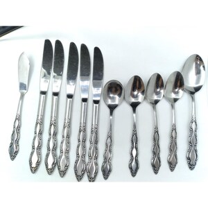 May include: A set of eleven stainless steel flatware pieces including five dinner knives, one butter knife, and five spoons. Each piece features a polished silver finish and a decorative embossed scroll pattern along the handle stems.