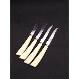 May include: Set of four steak knives with cream-colored handles and serrated stainless steel blades. The knives are arranged diagonally against a dark background, showcasing their elegant design and sharp edges.