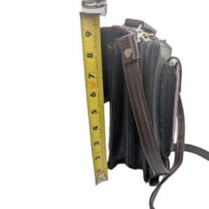 K&ouml;nnte beinhalten: Schwarze Aktentasche mit braunen Akzenten, mit Schultergurt und Griff. Die Aktentasche ist etwa 23 cm hoch, gemessen mit einem gelben Ma&szlig;band. Die Tasche hat ein strukturiertes Aussehen.