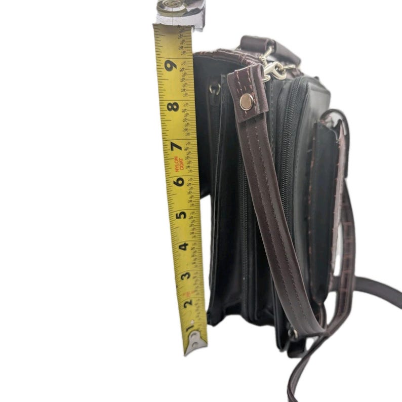 K&ouml;nnte beinhalten: Schwarze Aktentasche mit braunen Akzenten, mit Schultergurt und Griff. Die Aktentasche ist etwa 23 cm hoch, gemessen mit einem gelben Ma&szlig;band. Die Tasche hat ein strukturiertes Aussehen.