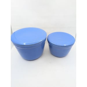 Op de afbeelding: Twee lichtblauwe keramische kommen met bijpassende deksels. De kommen hebben een gladde, glanzende afwerking en een taps toelopende, ronde vorm. De grotere kom staat links en de kleinere kom staat rechts, beide uitgevoerd in een effen kleur.
