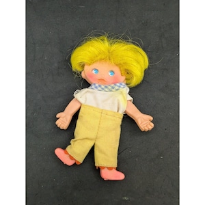 Puede incluir: Una muñeca con el pelo amarillo, ojos azules y un babero a cuadros, que lleva una camisa blanca y pantalones amarillos.