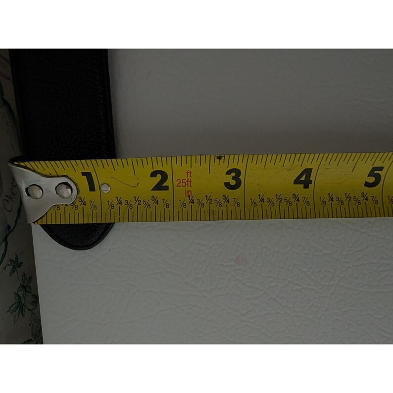 May include: A yellow measuring tape with black markings, showing measurements in inches. The tape is extended over a black object, with the measurement reading approximately 2 inches. The tape is marked with the text "25ft 10 in".