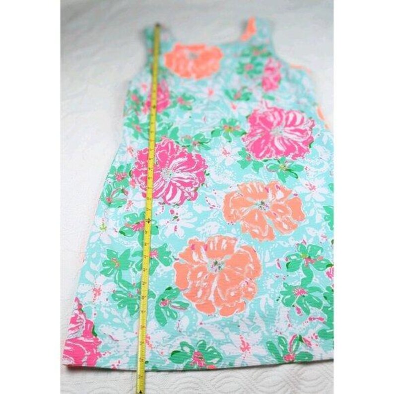 May include: A sleeveless dress with a floral pattern. The dress features large pink and orange flowers on a light blue background. Green and white accents complete the design. A yellow measuring tape is placed next to the dress.
