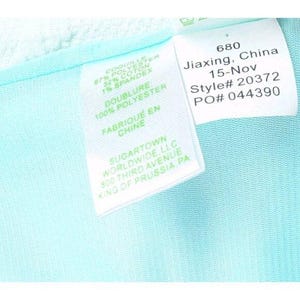 May include: Close-up of a white tag with black text on a light blue fabric. The tag includes details such as "67% POLYESTER, 32% COTON, 1% SPANDEX" and "FABRIQUE EN CHINE". Additional text includes "SUGARTOWN WORLDWIDE, LLC" and a style number.