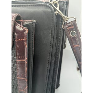 K&ouml;nnte beinhalten: Eine schwarze Lederaktentasche mit einem braunen, strukturierten Akzent. Die Aktentasche hat einen Rei&szlig;verschluss und einen abnehmbaren braunen Riemen mit einem silbernen Verschluss. Der Akzent hat ein gewebtes Muster.