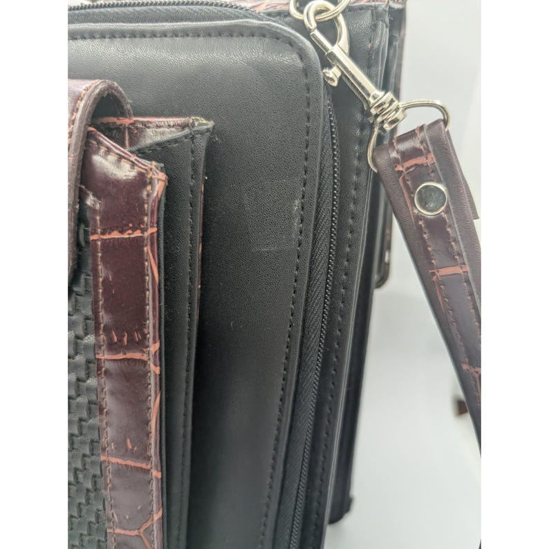 K&ouml;nnte beinhalten: Eine schwarze Lederaktentasche mit einem braunen, strukturierten Akzent. Die Aktentasche hat einen Rei&szlig;verschluss und einen abnehmbaren braunen Riemen mit einem silbernen Verschluss. Der Akzent hat ein gewebtes Muster.