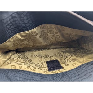 May include: The inside of a black leather handbag with a gold and brown patterned lining. The lining has a swirl design and the brand name is visible on a brown leather tag.