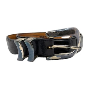 Könnte beinhalten: Schwarzer Ledergürtel mit silberner Schnalle und zwei silbernen Schlaufen. Der Gürtel hat ein strukturiertes Muster und eine goldfarbene Innenseite. Schnalle und Schlaufen weisen Gebrauchsspuren auf.