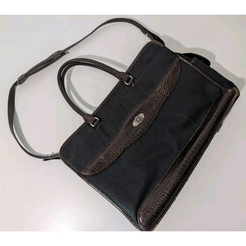 May include: A black leather briefcase with a brown crocodile embossed trim and a silver buckle. The bag has a top handle and a shoulder strap.