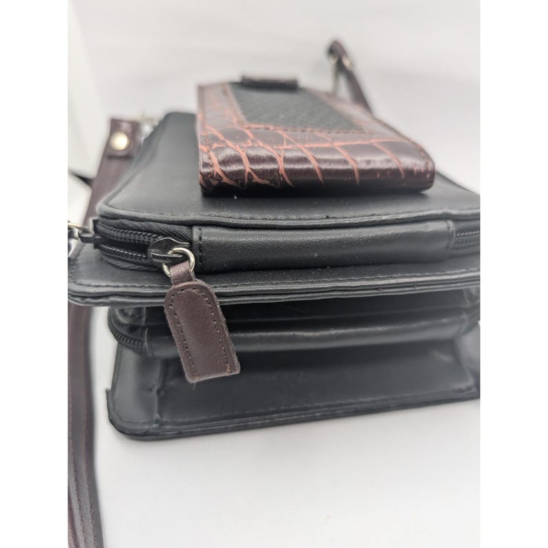 K&ouml;nnte beinhalten: Schwarze Umh&auml;ngetasche mit einer braunen, strukturierten Handyhalterung oben. Die Tasche hat ein Rei&szlig;verschlussfach und einen braunen Leder-Rei&szlig;verschlussanh&auml;nger. Ein brauner Riemen ist sichtbar.