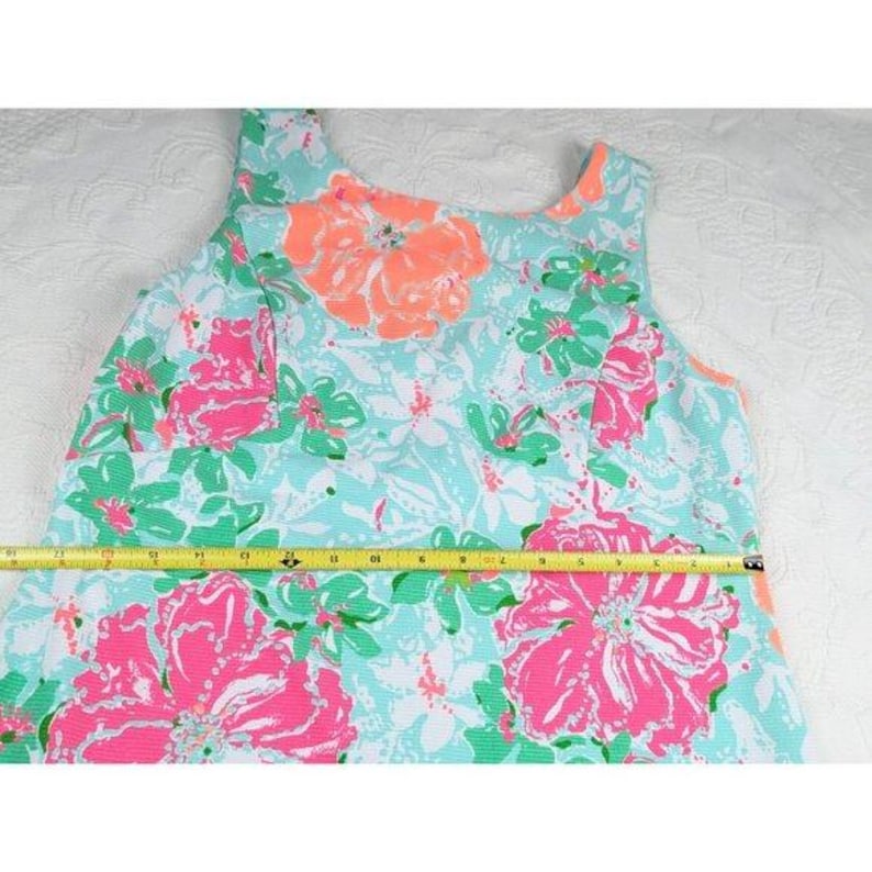 May include: A sleeveless dress with a floral pattern. The dress features large pink and orange flowers on a light blue background. Green leaves and smaller flowers are also present. A yellow measuring tape is laid across the dress.
