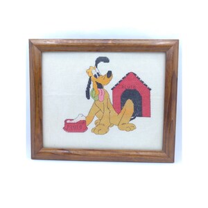 May include: A framed cross-stitch featuring Pluto, the Disney dog, sitting next to his red doghouse. The cross-stitch is on white fabric and the frame is brown wood.