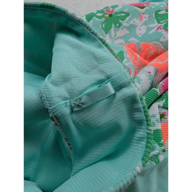 May include: Close-up of a turquoise fabric with a floral pattern in shades of green, pink, and orange. The fabric has a textured appearance and a small, attached ribbon detail. The overall design suggests a garment or textile.