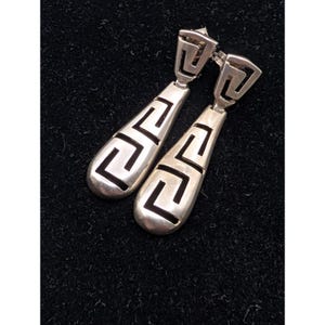 May include: A pair of silver drop earrings featuring a polished metallic finish and a cutout Greek key pattern. The earrings consist of a triangular top section connected to a longer, teardrop-shaped base, both displaying the same geometric motif.