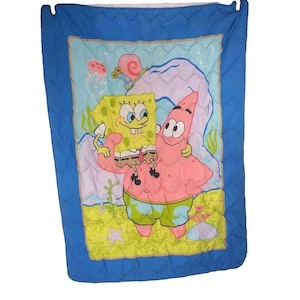 May include: A blue and yellow quilted blanket featuring SpongeBob SquarePants and Patrick Star. The characters are smiling and standing on a yellow sandy beach with a blue ocean background.