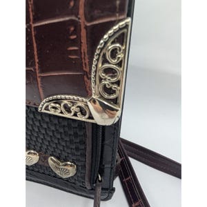 K&ouml;nnte beinhalten: Braune und schwarze Umh&auml;ngetasche mit strukturiertem, krokodil&auml;hnlichem Muster. Die Tasche hat einen gewebten schwarzen Bereich und dekorative silberfarbene Metallakzente. Ein brauner Riemen und Rei&szlig;verschluss sind sichtbar.