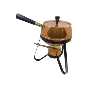 Op de afbeelding: Een koperen fonduepan met een zwarte handgreep en deksel. De pan staat op een zwarte metalen standaard met een kleine brander eronder. Een houten roerstaafje rust op de brander. De pan heeft een diameter van ongeveer 15 cm.