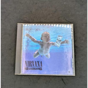 May include: A square compact disc case featuring the album cover for Nirvana Nevermind. The artwork displays a baby swimming underwater towards a dollar bill on a fishhook. The text Nirvana Nevermind is printed in a black serif font at the bottom.