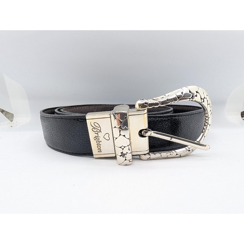 May include: Black leather belt with a silver-tone buckle. The buckle features a decorative design and the word "Brighton" is engraved on the buckle. The belt is rolled up on a white surface.