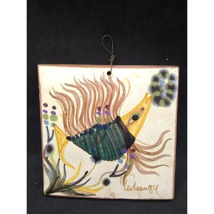 May include: A hand-painted ceramic tile featuring a stylized fish with yellow, green, and brown details. The fish is surrounded by stylized plants and flowers. The tile is signed "Wilstanzky" in the bottom right corner.