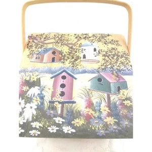 May include: A wooden box with a handle, painted with a whimsical scene of birdhouses in a field of flowers. The birdhouses are painted in various colors, including pink, green, and blue. The flowers are painted in shades of yellow, white, and purple.