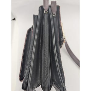 K&ouml;nnte beinhalten: Schwarze und braune Ledergeldb&ouml;rse mit mehreren Rei&szlig;verschlussf&auml;chern und einem Schulterriemen. Die Geldb&ouml;rse hat ein strukturiertes Muster auf der Au&szlig;enseite und einen goldfarbenen Verschluss. Der Innenraum ist schwarz.