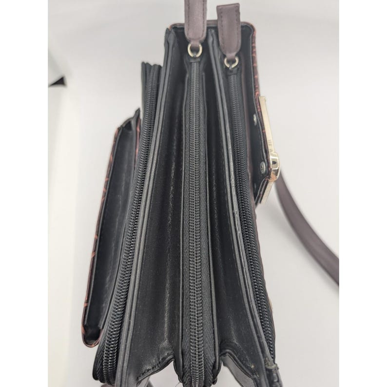 K&ouml;nnte beinhalten: Schwarze und braune Ledergeldb&ouml;rse mit mehreren Rei&szlig;verschlussf&auml;chern und einem Schulterriemen. Die Geldb&ouml;rse hat ein strukturiertes Muster auf der Au&szlig;enseite und einen goldfarbenen Verschluss. Der Innenraum ist schwarz.