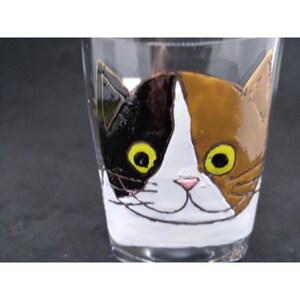 Puede incluir: Una taza de vidrio con una imagen pintada de la cara de un gato calico. El gato tiene pelaje negro, blanco y marrón con ojos amarillos.