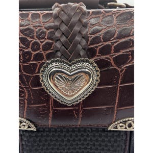 K&ouml;nnte beinhalten: Nahaufnahme einer braunen Lederhandtasche mit Krokodilmuster. Die Tasche hat einen geflochtenen Lederriemen und einen silbernen herzf&ouml;rmigen Verschluss. Der untere Teil der Tasche ist schwarz mit einer gewebten Textur und silbernen Akzenten.