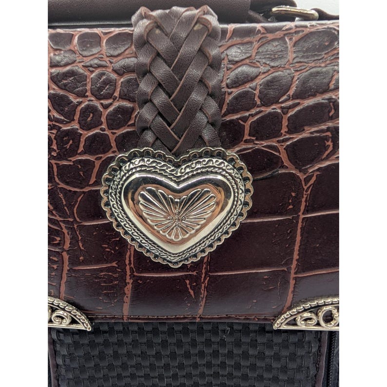 K&ouml;nnte beinhalten: Nahaufnahme einer braunen Lederhandtasche mit Krokodilmuster. Die Tasche hat einen geflochtenen Lederriemen und einen silbernen herzf&ouml;rmigen Verschluss. Der untere Teil der Tasche ist schwarz mit einer gewebten Textur und silbernen Akzenten.