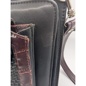 K&ouml;nnte beinhalten: Nahaufnahme einer schwarzen Ledergeldb&ouml;rse mit einem braunen, strukturierten Akzent. Die Geldb&ouml;rse hat einen Rei&szlig;verschluss und einen braunen Lederriemen mit einem silberfarbenen Verschluss. Der Akzent hat ein gewebtes Muster.