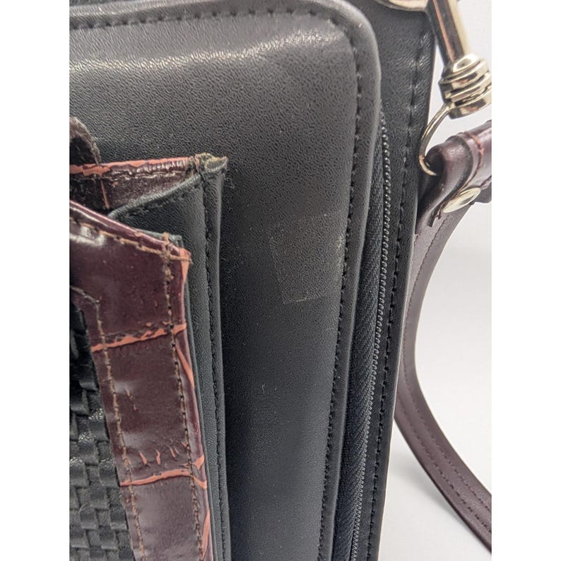 K&ouml;nnte beinhalten: Nahaufnahme einer schwarzen Ledergeldb&ouml;rse mit einem braunen, strukturierten Akzent. Die Geldb&ouml;rse hat einen Rei&szlig;verschluss und einen braunen Lederriemen mit einem silberfarbenen Verschluss. Der Akzent hat ein gewebtes Muster.